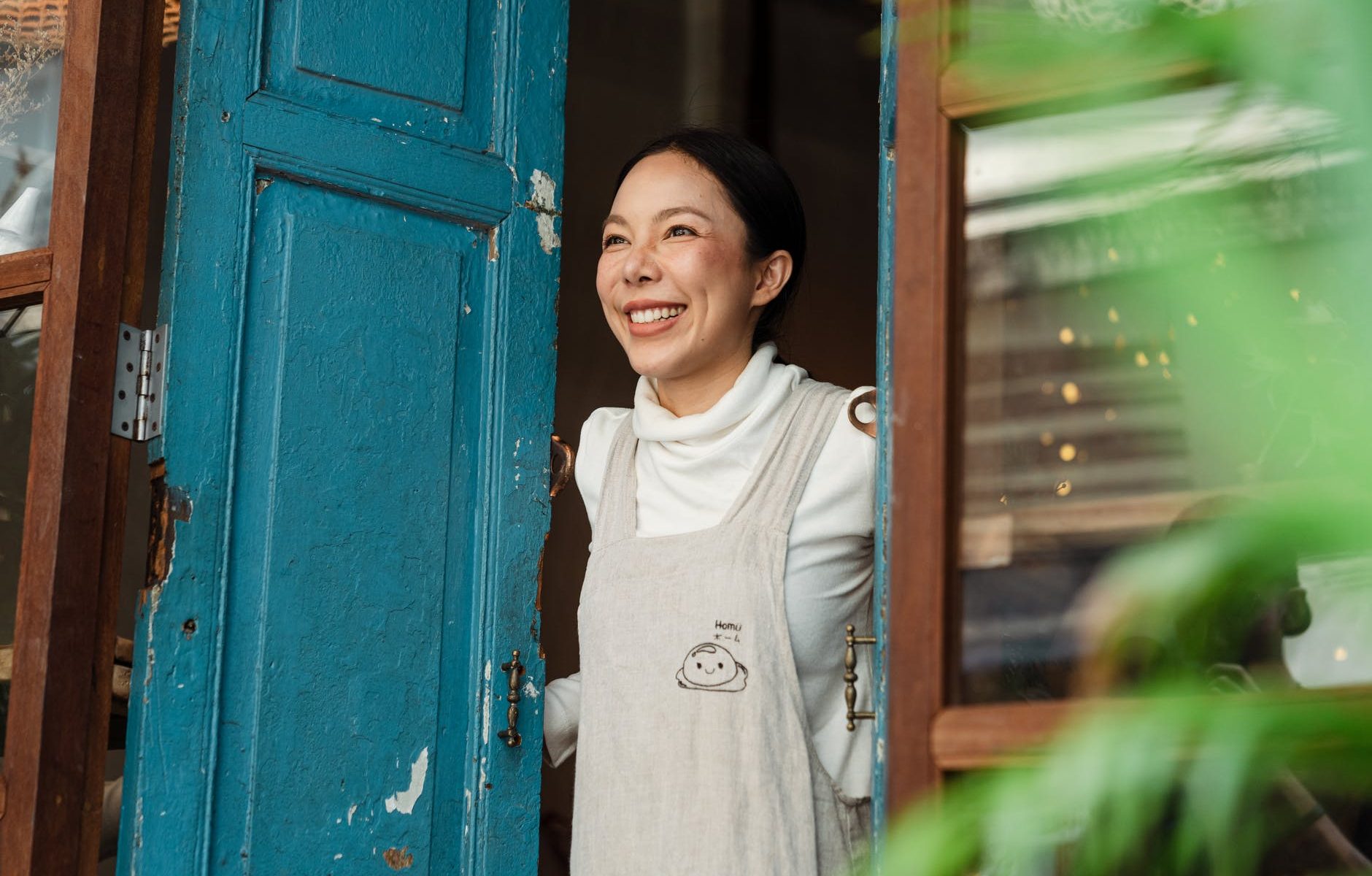 happy ethnic woman in apron standing at entrance of own cafe